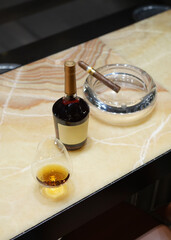 Close-up of a snifter glass filled with cognac, a cigar resting on a glass ashtray, and cognac bottle on a marble bar counter. Luxury and relaxation concept