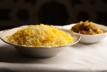 Fluffy basmati rice infused with saffron, displayed in a decorative white bowl. A stew or curry is visible in a slightly blurred bowl in the background