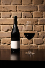 A bottle of red wine with a blank label and a glass of red wine, standing on a dark wooden table against a brick wall background with copy space