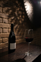 A bottle of red wine with a blank label and a glass of red wine, standing on a dark wooden table against a brick wall background with copy space
