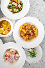 A top-down view of a diverse spread of plates featuring various dishes, including salads, pasta, and a colorful seafood dish, set on a textured light grey background.