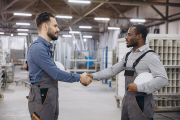 Two interracial factory workers shaking hands in a metalwork carpentry, they're both holding safety helmets