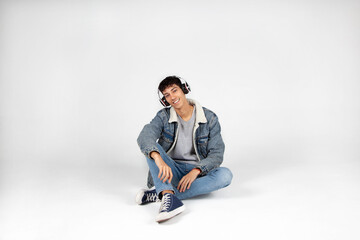 Chico joven vestido de vaqueros y camiseta blanca escuchando música con auriculares, sonriendo a cámara. Fondo blanco. Concepto de música, estilo casual y publicidad.