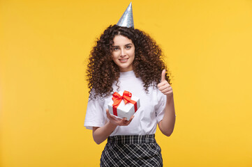 Smiling young woman in party hat showing thumbs up while holding a present box on yellow...
