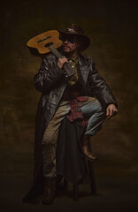 Brutal man, cowboy sitting on stool in leather coat, hat and boots, holding acoustic guitar over shoulder, looking away against dark studio background. Concept of music, western lifestyle, history.