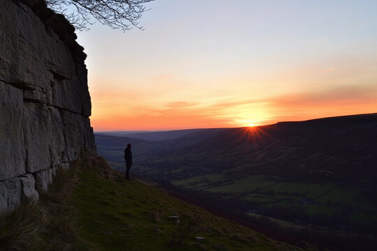 Silhouette of a person standing at the edge of a cliff during sunset in a mountainous landscape