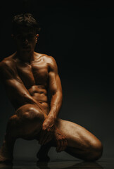 Fototapeta premium A strong and toned man showcases his physique in a dramatic low-light studio setting, highlighting shadows and textures for an artistic, elegant representation of the human body.