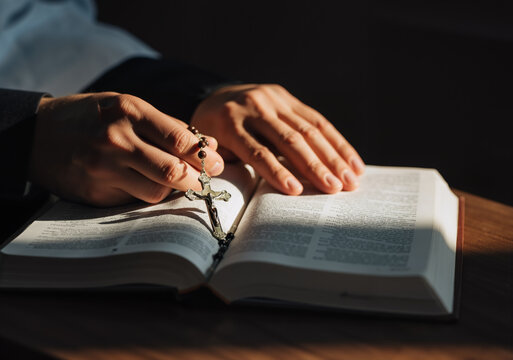 Priest is holding rosary beads and praying with the open holy bible on a wooden table in a dark room with a ray of light
