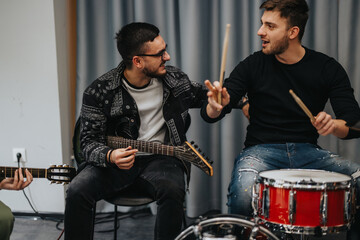 A group of friends playing instruments and engaging in music practice in a relaxed studio atmosphere, focusing on drums and guitars while bonding creatively over music.