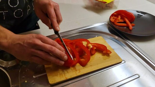 Close-up of a left-handed man slicing red bell peppers on a wooden cutting plate in his home kitchen