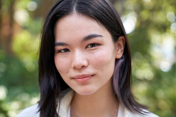 Fototapeta premium Headshot of a serene young japanese woman with dark hair and brown eyes, wearing a white shirt, posing in a park with a blurred background