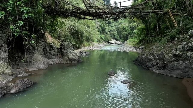 Drone flying low over the calm flowing river water, passing under the root bridge as an exotic icon of the Baduy tribe and as a cultural heritage tourism destination in Banten, Indonesia.