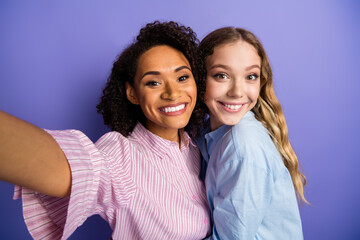Two young women smiling brightly in a vibrant selfie against a purple background, showcasing friendship