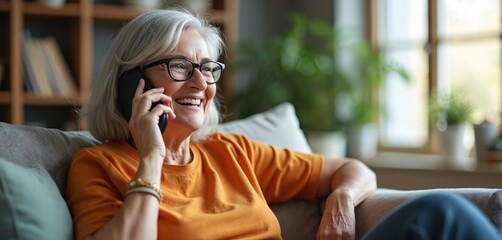 Happy mature woman wearing glasses talking with relatives on mobile phone sitting on sofa at home. Senior citizen shares news, advice, listens. Grandmother communicates on smartphone, modern mobile