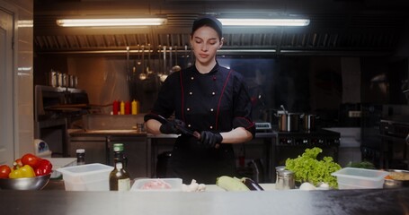 A woman in an apron and cap sharpens knives standing in an industrial kitchen, preparing for cooking