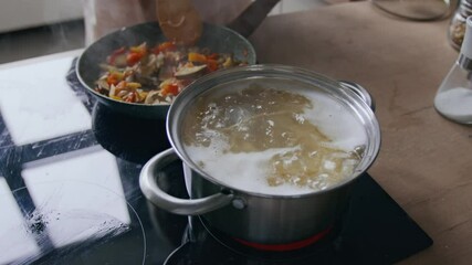 Closeup of unrecognizable person frying vegetables in pan, stirring them with wooden spoon, while spaghetti boiling nearby on modern cooktop in bright, cozy kitchen setting - Powered by Adobe