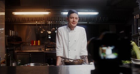A man in a chef's uniform records the cooking process on video, standing near a frying pan in the kitchen of a restaurant
