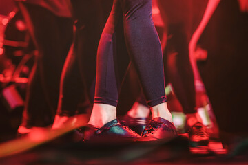 Feet of dancers in black shoes, energetic dance steps on stage, red lighting, spirit of ethnic dance. Bright blurred lighting stage background