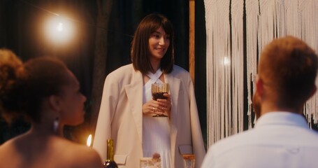 Guests smile sitting at a beautifully served festive table outdoors at night under the light bulbs, a woman stands saying congratulations