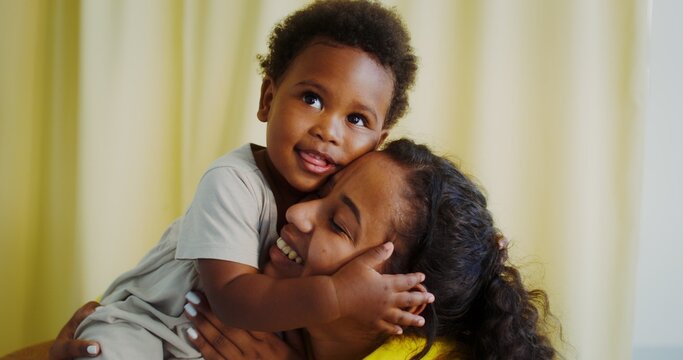 Mom and a baby of African-American appearance hug and smile. The child is sitting on a chair, and the mother is on the floor next to her