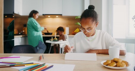 A girl draws with a stylus on a tablet while sitting at the kitchen table at home. Her family in...