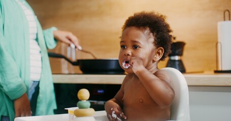 A baby of African-American appearance is eating yogurt with a spoon sitting in a chair, drops of...