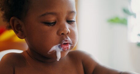 A baby of African-American appearance is eating yogurt with a spoon sitting in a chair, drops of...