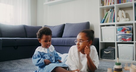 A teenager and a baby girl of African-American appearance watch cartoons on a tablet while sitting on the floor at home