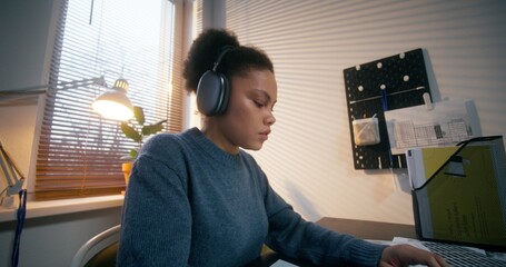 A woman in casual clothes, wearing headphones, typing on a laptop sitting at a desk working in an...