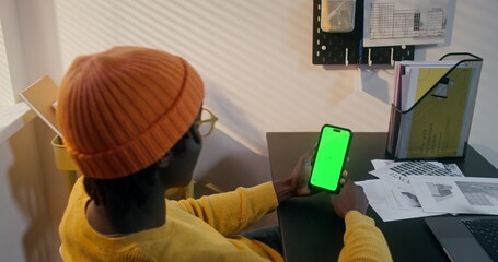 A man in informal clothes uses a mobile phone with green screen sitting at a desk in the office