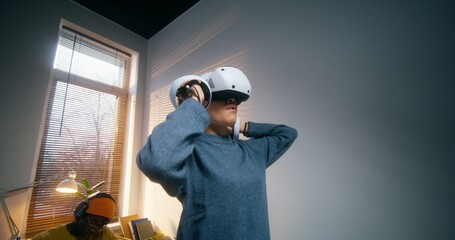 A young woman in a virtual reality headset uses a joystick standing in an office. A man draws on a...
