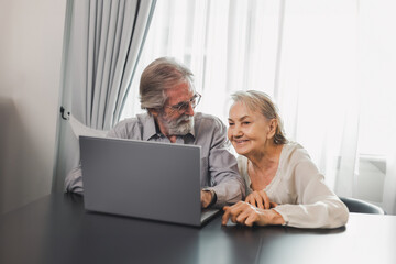 Happy Caucasian 70s mature couple using laptop at home. Old people using computer at home. Senior man and woman surfing the web on a wireless laptop connection. Modern retired lifestyle people.