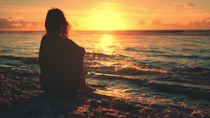A woman is seated on the sandy beach, gazing out at the horizon where the sun is setting. The sky displays vibrant hues of orange, pink, and purple as the sun ly disappears below the water.