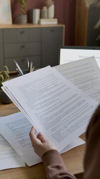Vertical closeup of hands of anonymous woman sitting at desk at home, studying draft of mortgage agreement on laptop screen and paper copy, while planning to buy new house