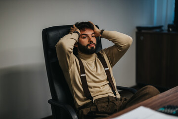 Obraz premium A contemplative professional man is seated at his desk, hands resting behind his head. The office background creates a setting suggestive of introspection, business challenges, and potential
