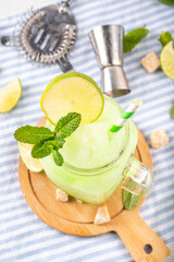 Mojito lemonade alcoholic slush cocktail, garnished with mint and lime slice, summer refreshing slushy drink with vodka, lime juice and mint syrup, on a white table with bar utensils