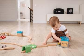 A two-year-old boy in a brown overall and white T-shirt plays with toys at home, enjoying a fun and...