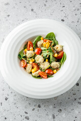A vibrant salad featuring sesame-coated bites, fresh spinach, cherry tomatoes, and sweet corn, elegantly arranged in a white bowl on a textured surface.