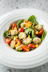 A vibrant salad featuring sesame-coated bites, fresh spinach, cherry tomatoes, and sweet corn, elegantly arranged in a white bowl on a textured surface.