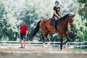 A young rider on a brown horse is observed by a trainer during practice in an outdoor arena. The...