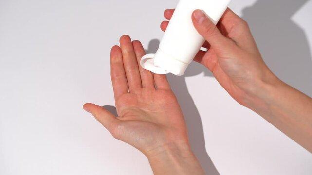 Female hands open white mockup tube of cream lotion and apply it on palm of her hand rubbing it on white isolated table background. Concept of skin nourishment and hydration, body care, beauty product