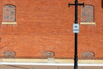 full frame red brick wall with bricked in blind windows and view of sidewalk urban background frame with space for copy. a black and white no loitering sign is affixed to a metal lamppost by pavement.