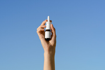 Young woman's hand sprays nasal spray from a glass mockup on a blue sky background isolated. Concept of nasal cleansing, clean breathing during flu and illness