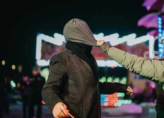 Two friends are playfully interacting with each other under colorful lights at a carnival, creating...