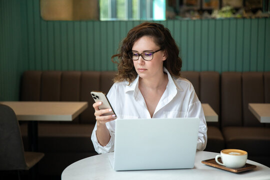Young happy woman sitting at table holding smartphone using cellphone modern technology, looking at mobile phone while remote working or learning, texting messages at home Tech, mobile app concept
