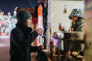 Two friends smiling and enjoying hot drinks at a seasonal market on a chilly evening.