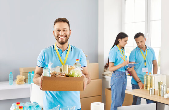 Portrait of a happy Caucasian man volunteer holding a box of food and looking at camera in charity organization. Contribution to donations and service in helping others through charitable activities.
