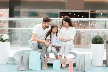 Family, father, mother and daughter are sitting on bench in shopping mall. Girl is looking inside one bag.
