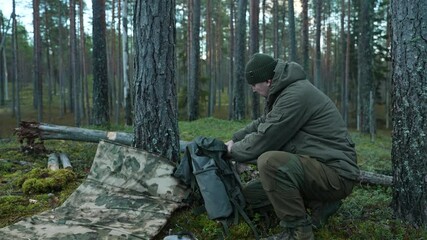 A man is packing his gear into a camping backpack in the forest. A bushcrafter is organizing his backpack in a coniferous forest among the trees. The concept of survival in the wild