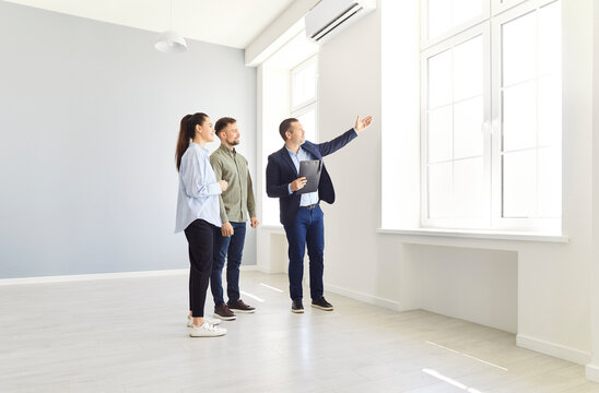 Fototapeta Mature real estate agent, realtor standing with potential buyers in new house or apartment, showing them amazing view from window. Young family couple standing in empty room, buying new house.